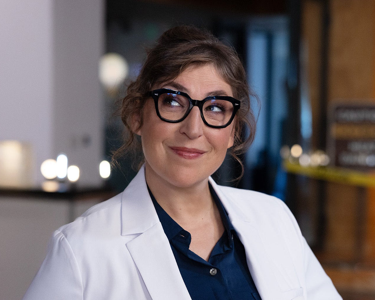 Mayim Bialik wearing a white cree lighting lab coat and glasses in an indoor