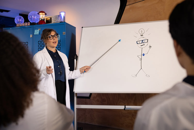 Mayim Bialik in a white cree lighting lab coat standing next to a whiteboard 